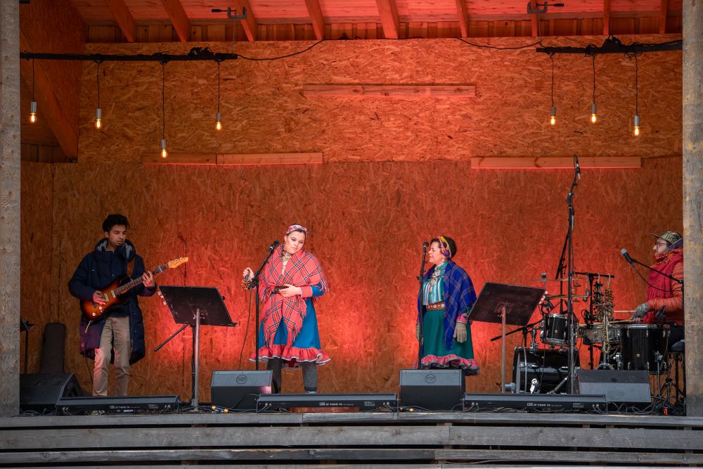 Kaksi saamelaisten kansallispukuihin pukeutunutta naista laulaa esiintymislavalla, jossa on myös kitaristi ja rumpali