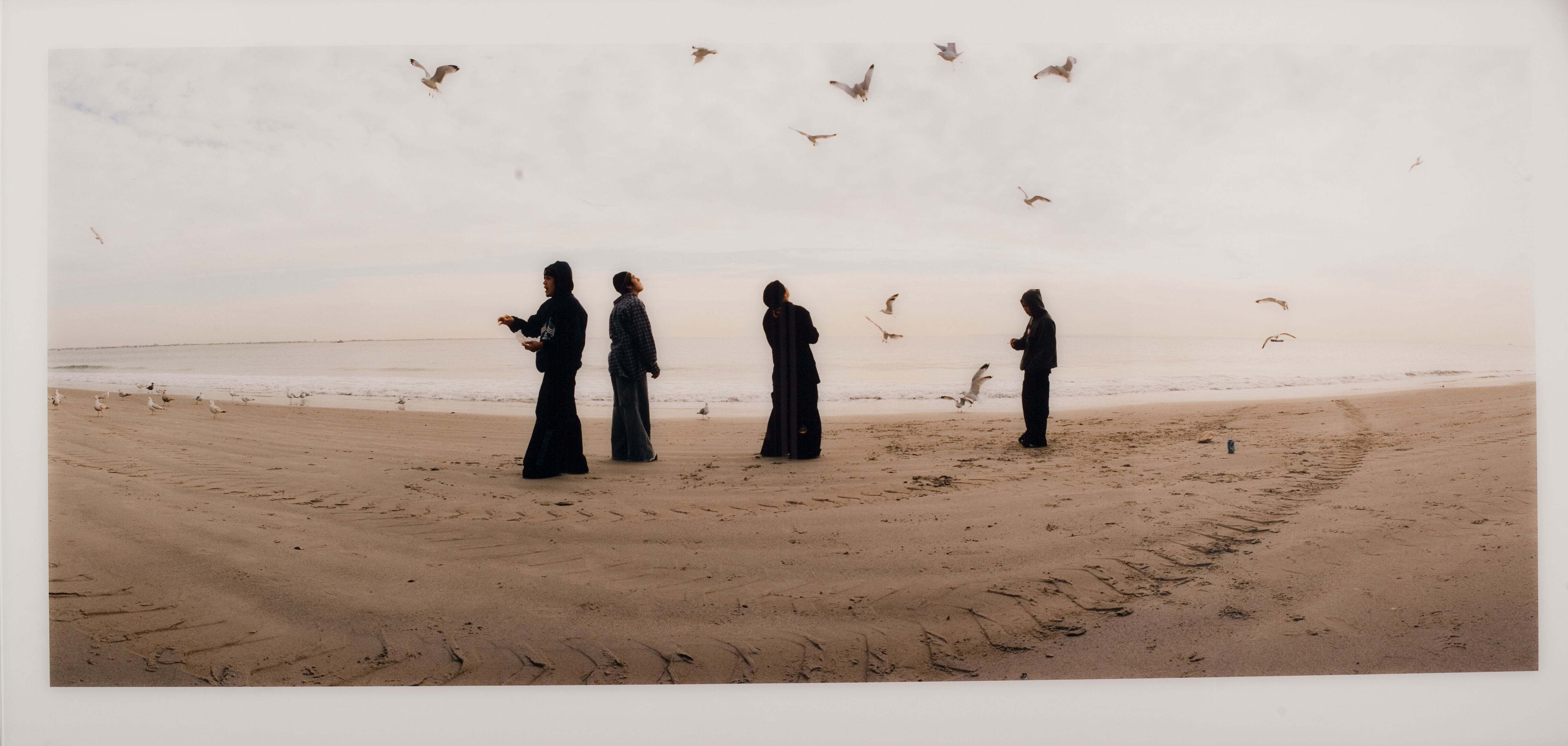 Neljä mustiin pukeutunutta henkilöä rannalla. Taivaalla lentää lintuja.