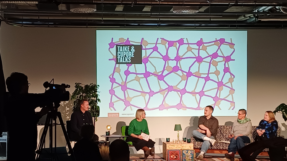 Taike & Cupore -talks-keskustelijat lavalla. Reunassa videokameran ja kuvaajan siluetti.