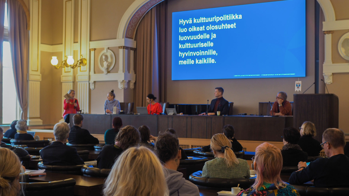 Suuren salin lavalla viisi ihmistä ja taustalla valkokankaalla teksti "Hyvä kulttuuripolitiikka luo oikeat olosuhteet luovuudelle ja kulttuuriselle hyvinvoinnille, meille kaikille."