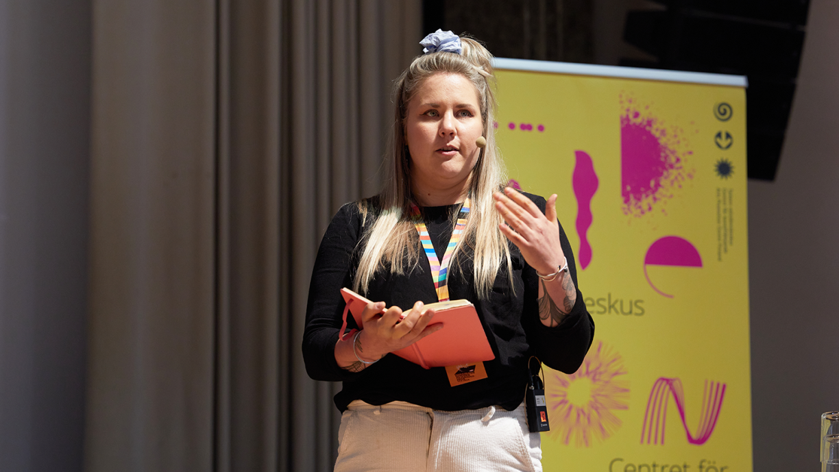 Suvi Sysi puhumassa lavalla. Taustalla Taiken Visuaalisten taiteiden tulevaisuuspäivän bannerolli.