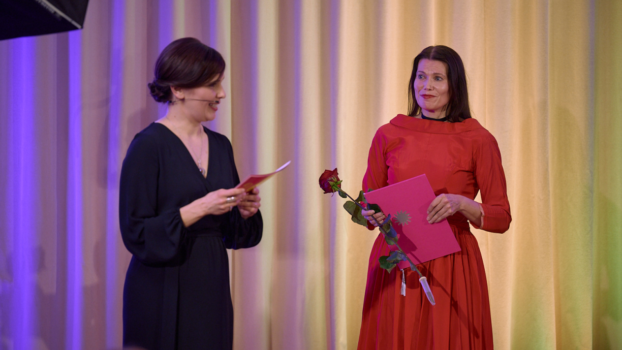 Kaksi henkilöä lavalla, joista toinen lukee kortista ja toinen kuuntelee.