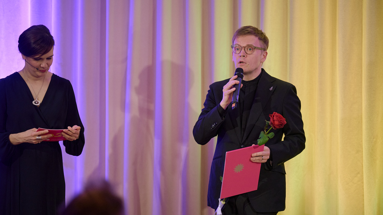 Pekka Kuusisto puhuu mikrofoniin kädessään kunniakirja ja ruusu.