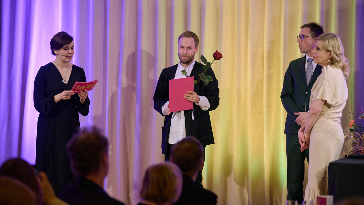 Ihmisiä lavalla, joista yksi lukee ja muut kuuntelevat.