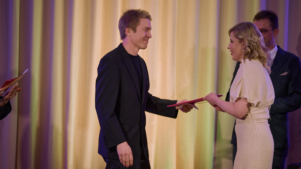 Henkilö ottaa kädellään vastaan kunnikirjan, jota toinen henkilö ojentaa.