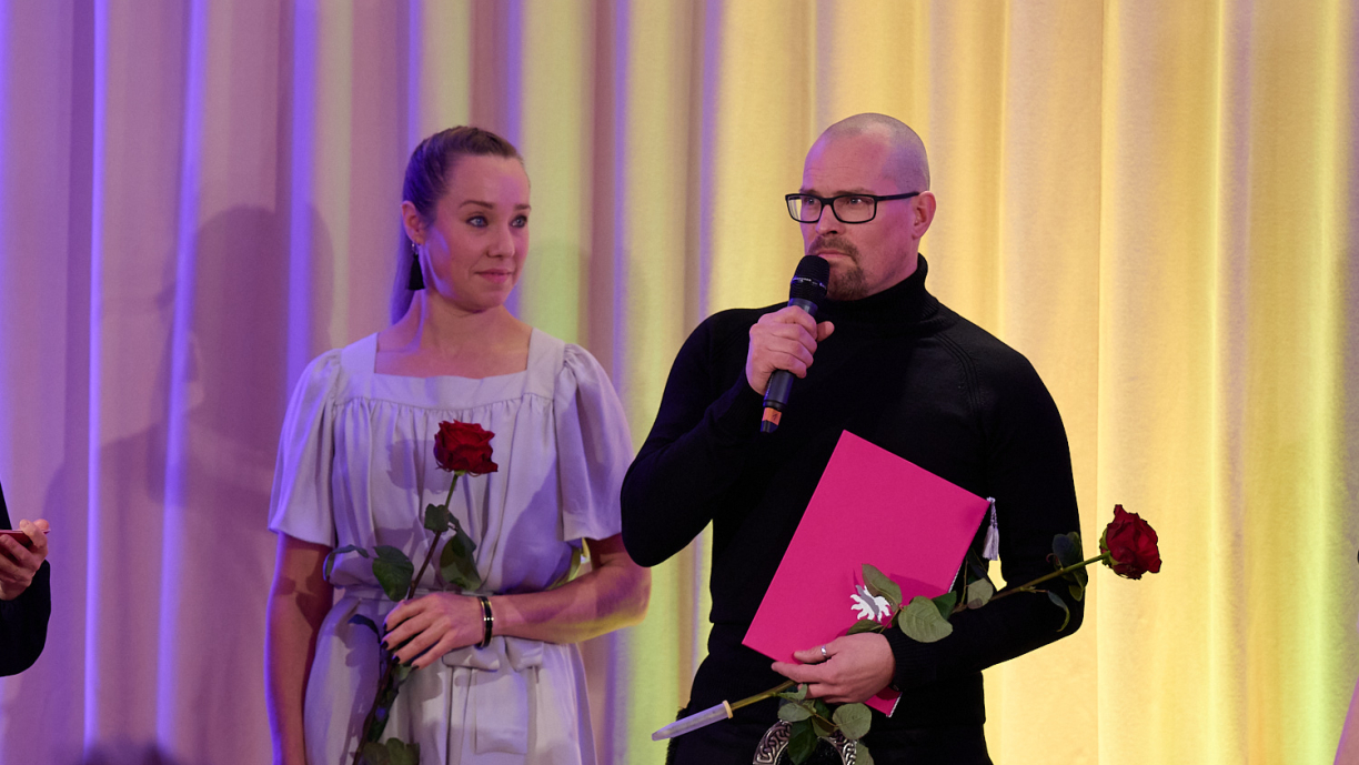 Kaksi henkilöä lavalla, joista toinen puhuu mikrofoniin.