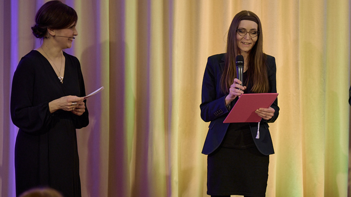 Kaksi henkilöä lavalla, joista toinen puhuu mikrofoniin.