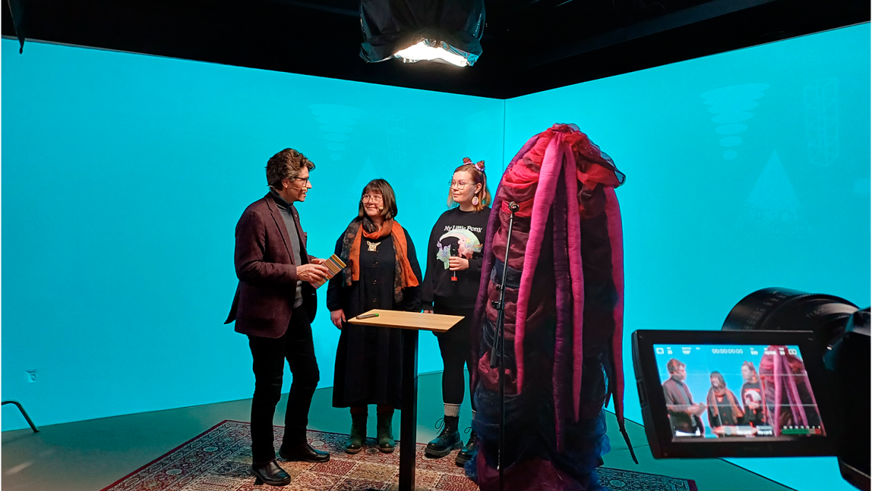 Neljä henkilöä seisovat studiossa pöydän ympärillä.