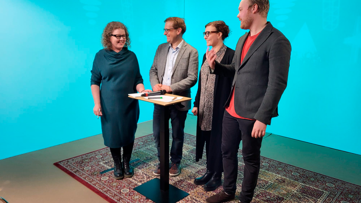Neljä henkilöä seisovat studiossa pöydän ympärillä.