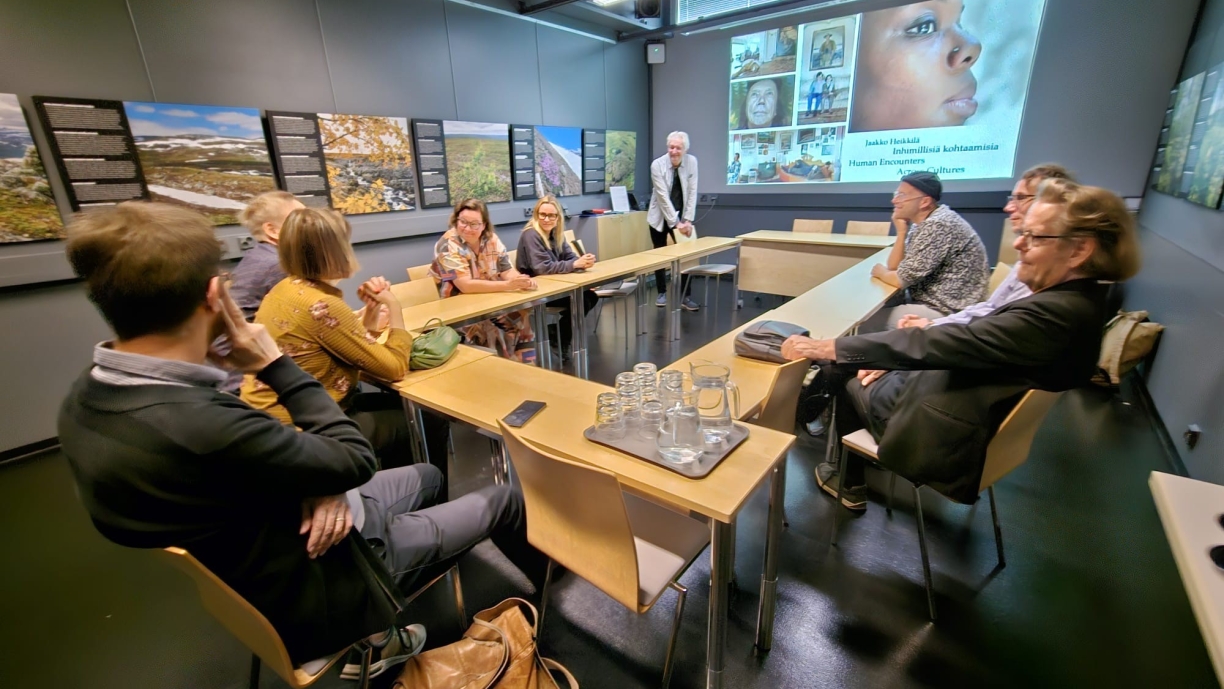 Ihmisiä istumassa pöydän ääressä. Seinälle heijastettuna esitys.