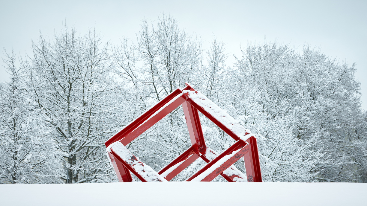 Punainen metallipalkeista tehty kuutio kulmittain lumisessa maassa.