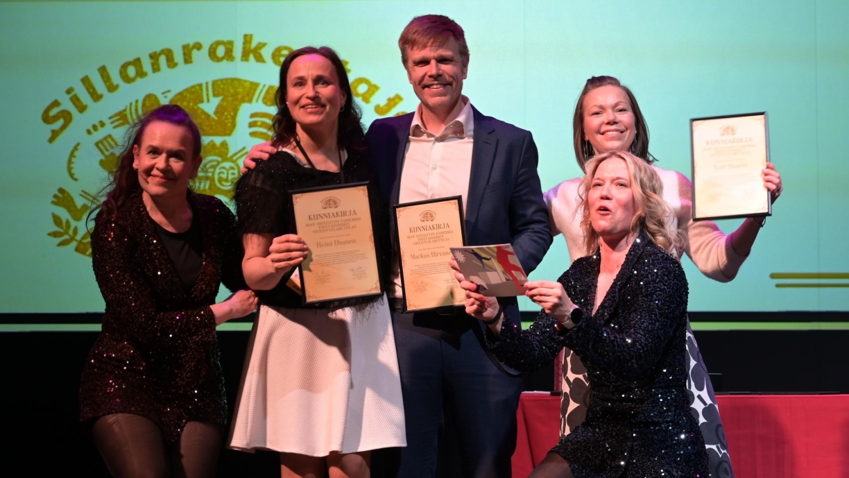 Maakuntajohtajat seisovat kunniakirjat käsissään. Sillanrakentajien Virpi ja Tiia-Mari ovat reunoilla glitter-mekoissa.