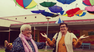Kaksi naista levittää käsiään ja näyttää hämmästyneeltä. Heidän yläpuoleltaan sataa sateenvarjoja.
