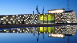 Valokuva Merikeskus Vellamosta pimeällä. Kuva otettu vastapäiseltä rannalta ja rakennuksen valot heijastavat rakennuksen kuvan tyynen veden pintaan. 