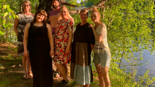 Kuusi ihmistä seisovat kesäisessä maisemassa puun edessä.