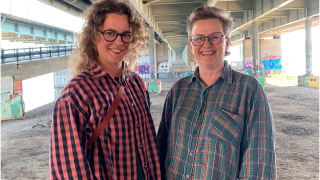 Kaksi hymyilevää ihmistä kuvattuna ison autotiesillan alla.