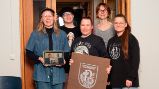 Viisi ihmistä seisoo muodostelmassa ja yhdellä on kädessään kyltti.