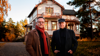 Kaksi henkilöä seisoo puutalon edessä.