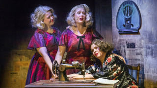 Kolme naista lavalla, joista yksi istuu ja kaksi seisovat ja keskimmäinen laulaa.