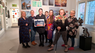 Seitsemän ihmistä seisoo galleriatilassa ja yksi pitää kylttiä kädessään.