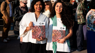 Kaksi henkilöä pitävät käsissään lehteä ja takana ihmisvilinää.