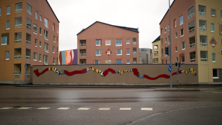 Teos kaupunkitilassa. Kerrostalon pihamuurissa luikertaa monivärinen muoto.