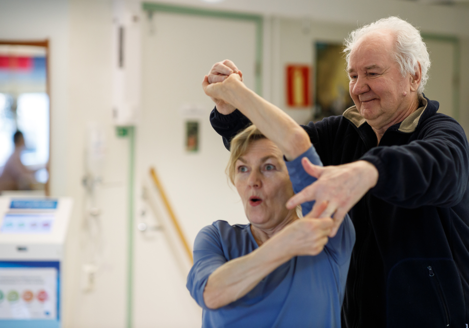 Två personer dansar tillsammans och håller varandra i händerna. En kvinnlig danskonstnär i förgrunden, med ett entusiastiskt ansiktsuttryck. Bakom henne en äldre, gråhårig man som ler och styr kvinnan med sina händer.
