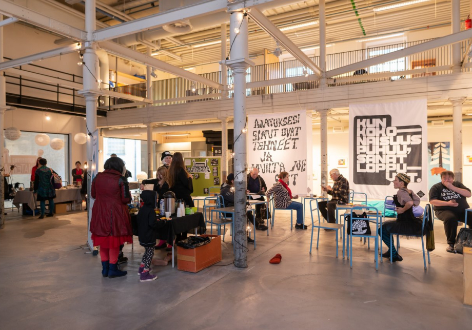 Ihmisiä istumassa tuoleilla ja seisomassa esittelypöydän ääressä kirjallisuuden esittelytilaisuudessa.