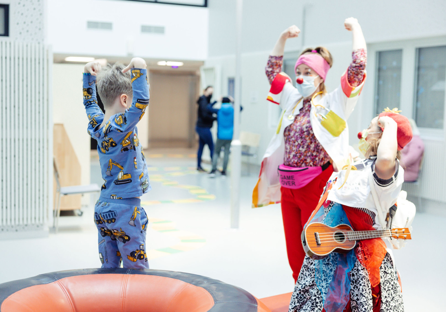 Kaksi sairaalaklovnia punaisissa nenissään leikkivät lapsipotilaan kanssa.