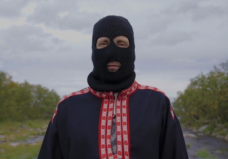 Sámi Hustler stands outdoors wearing a black balaclava on his head and looks directly at the camera.