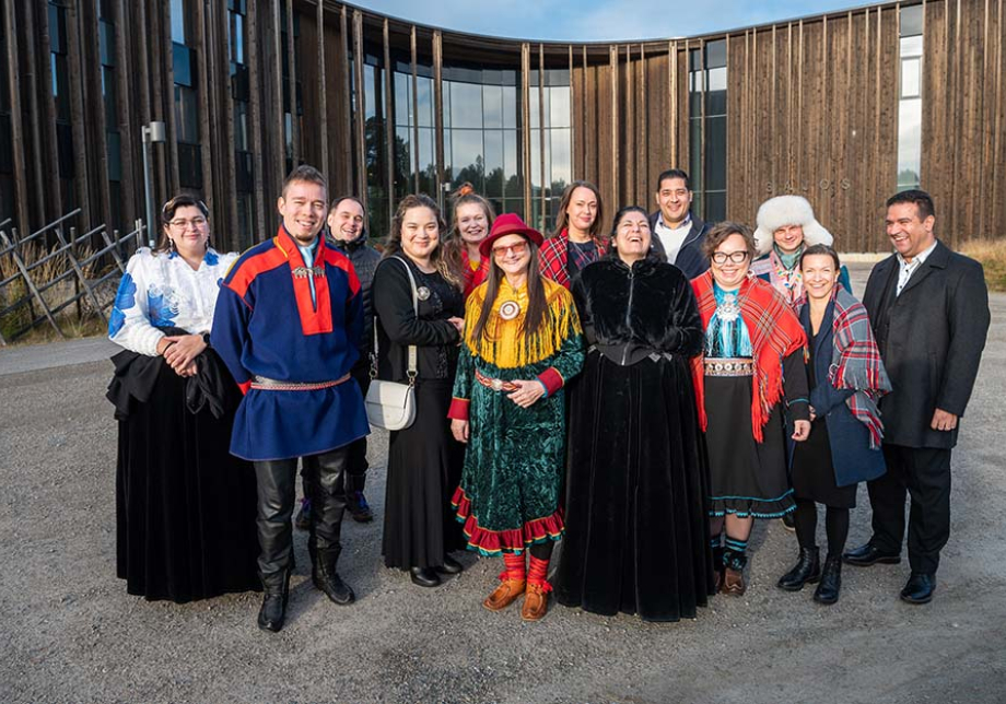 Joukko ihmisiä on kerääntynyt yhteen ja hymyilee kameralle. Taustalla on puuseinäinen rakennus.