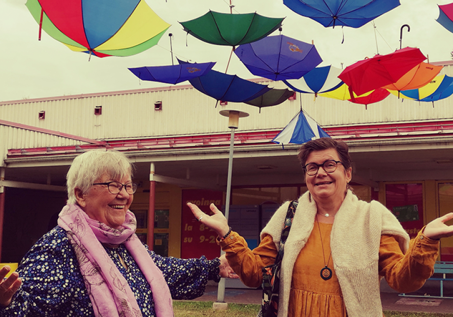 Kaksi naista levittää käsiään ja näyttää hämmästyneeltä. Heidän yläpuoleltaan sataa sateenvarjoja.