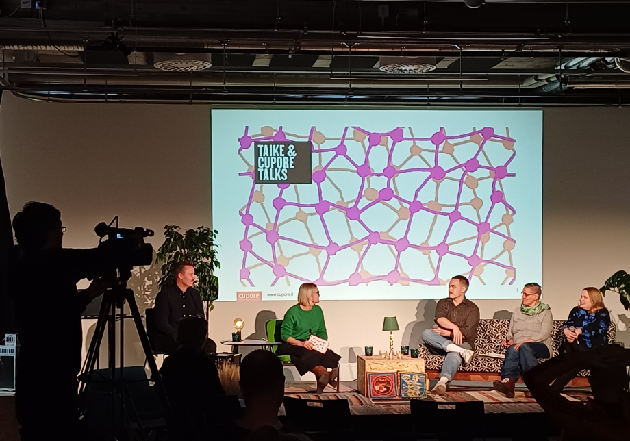 Taike & Cupore -talks-keskustelijat lavalla. Reunassa videokameran ja kuvaajan siluetti.