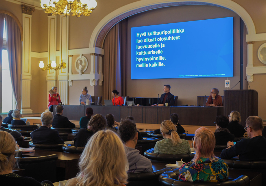 Suuren salin lavalla viisi ihmistä ja taustalla valkokankaalla teksti &quot;Hyvä kulttuuripolitiikka luo oikeat olosuhteet luovuudelle ja kulttuuriselle hyvinvoinnille, meille kaikille.&quot;