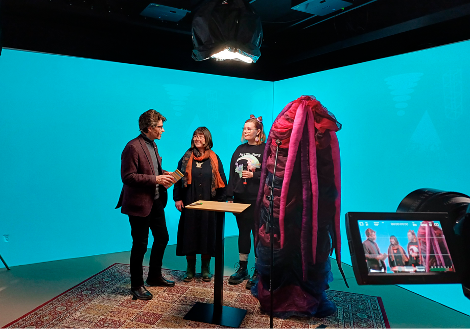 Neljä henkilöä seisovat studiossa pöydän ympärillä.