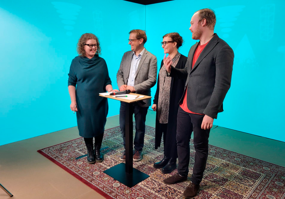 Neljä henkilöä seisovat studiossa pöydän ympärillä.