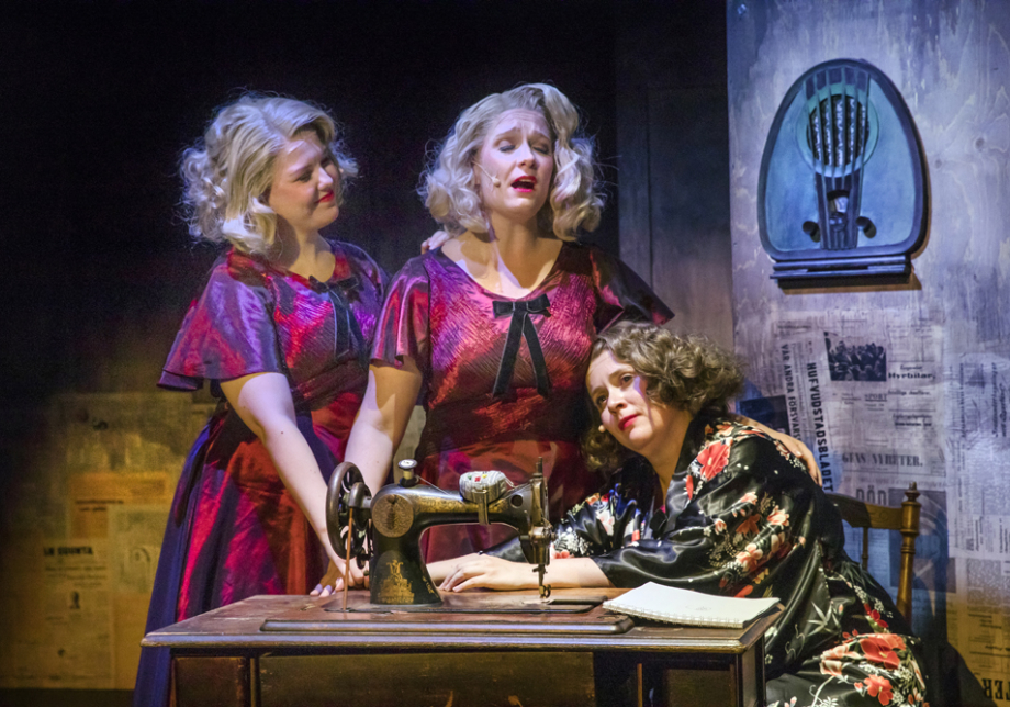 Kolme naista lavalla, joista yksi istuu ja kaksi seisovat ja keskimmäinen laulaa.