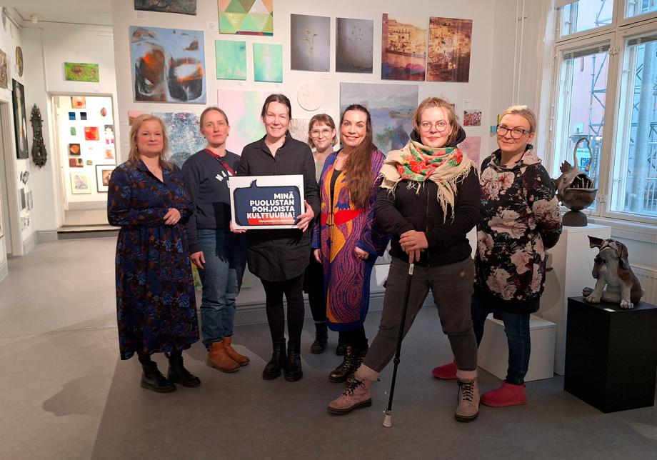 Seitsemän ihmistä seisoo galleriatilassa ja yksi pitää kylttiä kädessään.