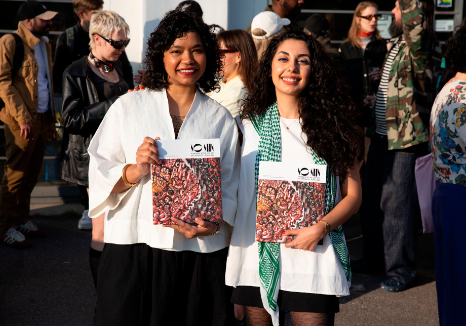 Kaksi henkilöä pitävät käsissään lehteä ja takana ihmisvilinää.