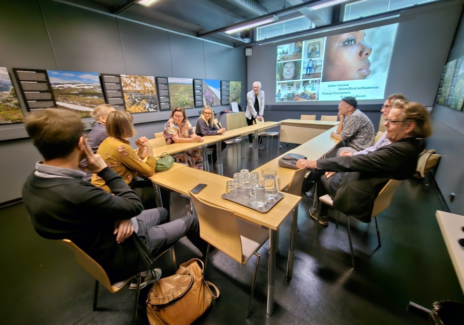 Ihmisiä istumassa pöydän ääressä. Seinälle heijastettuna esitys.