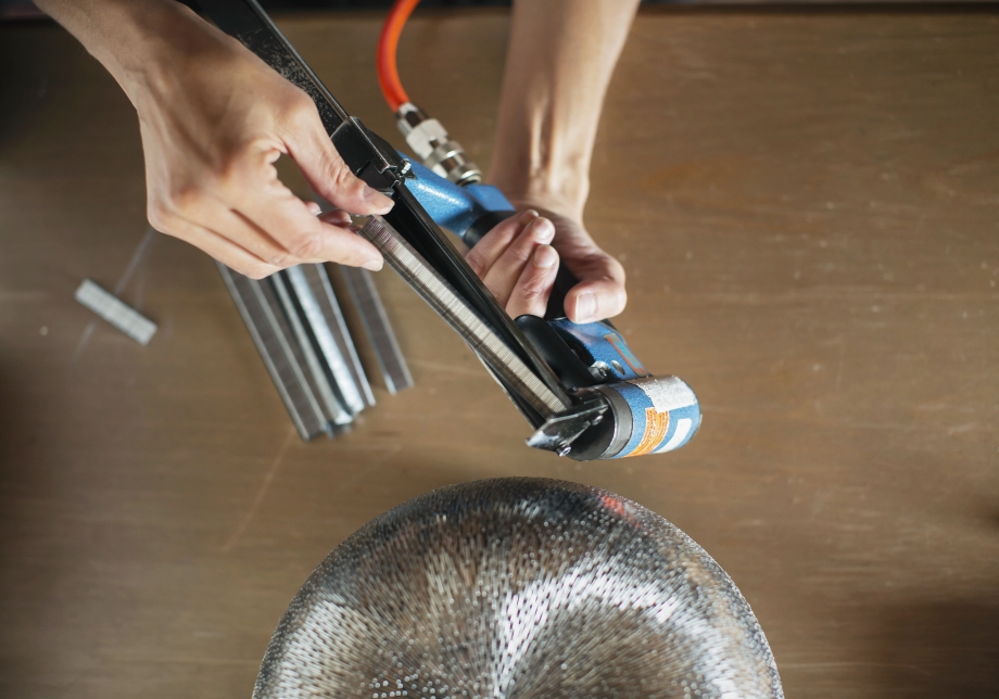 Taiteilijan käsissä on niittipyssy, jolla tekee metallinväristä niittiteosta