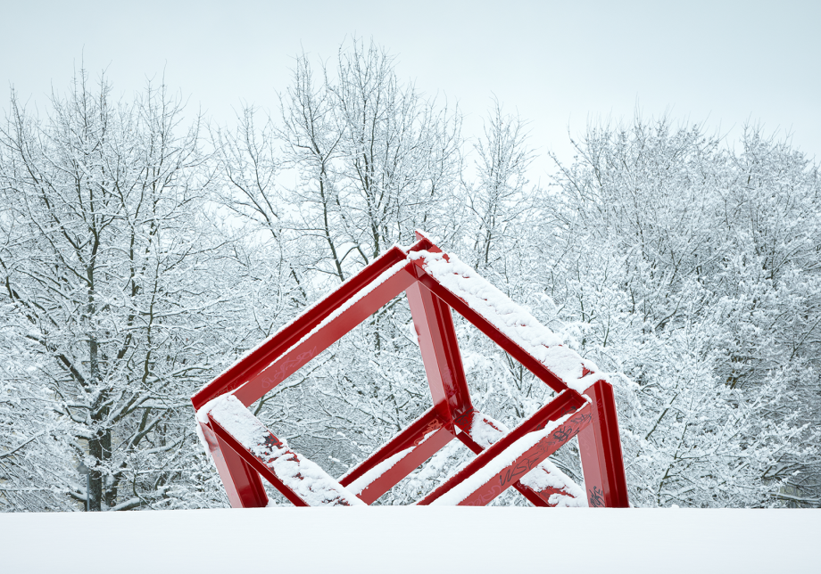 Punainen metallipalkeista tehty kuutio kulmittain lumisessa maassa.