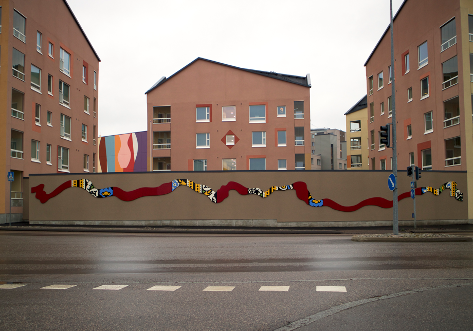Teos kaupunkitilassa. Kerrostalon pihamuurissa luikertaa monivärinen muoto.